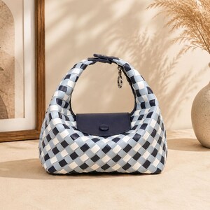 May include: A small handbag featuring a woven pattern in shades of navy blue, light blue, and white. The bag has a rounded top handle, a solid navy blue flap closure with a single matching button, and a small silver-toned metal ring attachment.