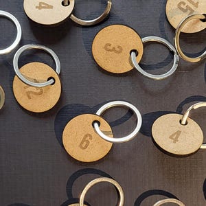 May include: A selection of numbered wooden keyrings with silver split rings. The keyrings are circular and feature engraved numbers from 1 to 15. The keyrings are arranged on a dark surface.