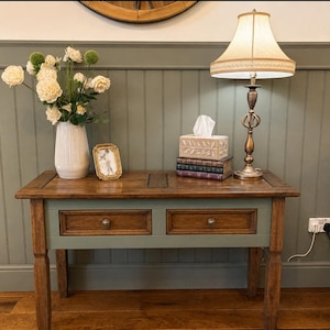 May include: A rectangular wooden console table with two drawers, featuring a natural wood top and a sage green base. On the table sits a white vase with white flowers, a small gold picture frame, a stack of books, a tissue box, and a table lamp.