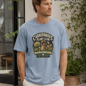 May include: A light blue t-shirt featuring a graphic print with the text "CARNIVOROUS PLANT CLUB", "TRAPS - TENDRILS - BOG LIFE", and "RARE LEAF SOCIETY" alongside illustrations of various carnivorous plants and a small skull icon.