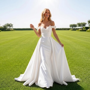 Peut inclure: Une robe de mariée blanche longue au sol avec une encolure dégagée ornée d'applications florales délicates. La robe présente un corsage ajusté au design drapé et une jupe volumineuse et fluide qui se prolonge en une longue traîne élégante.