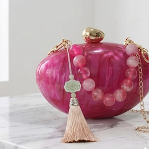 Peut inclure: Une pochette en acrylique marbré fuchsia avec un fermoir ovale doré. Le sac présente une anse décorative composée de perles roses translucides, une breloque géométrique vert clair et un pompon en tissu beige avec un embout en métal argenté.