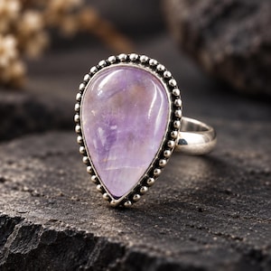Pode incluir: Um anel de prata com uma grande pedra preciosa roxa clara em forma de pera, engastada numa moldura com uma borda decorativa de contas. A banda de prata lisa e polida liga-se ao engaste que segura a pedra translúcida e lisa.