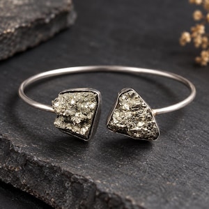 Puede incluir: Una pulsera abierta de tono plateado con dos piedras de pirita metálicas y en bruto en cada extremo. Las piedras tienen una textura cristalina facetada de color oro latón, engastadas en marcos de metal liso unidos a una banda fina.
