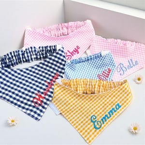 May include: A collection of personalized dog bandanas in various colors, including pink, blue, yellow, and navy, with a gingham pattern. Each bandana has a name embroidered in a contrasting color. The bandanas are arranged on a white surface.