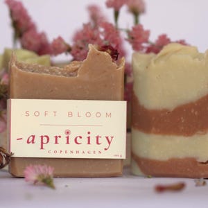 Könnte beinhalten: Zwei handgemachte Seifenstücke mit einem Blumengesteck. Ein Stück ist hellbraun mit einem Etikett mit der Aufschrift "SOFT BLOOM - apricity COPENHAGEN". Das andere Stück hat ein geschichtetes Design aus Weiß und Rotbraun. Rosa Blumen sind um die Seifen verstreut.