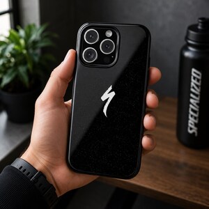 May include: Black phone case featuring a white Specialized logo. The case is held in a hand, with a black watch on the wrist. A black water bottle with "SPECIALIZED" is in the background.
