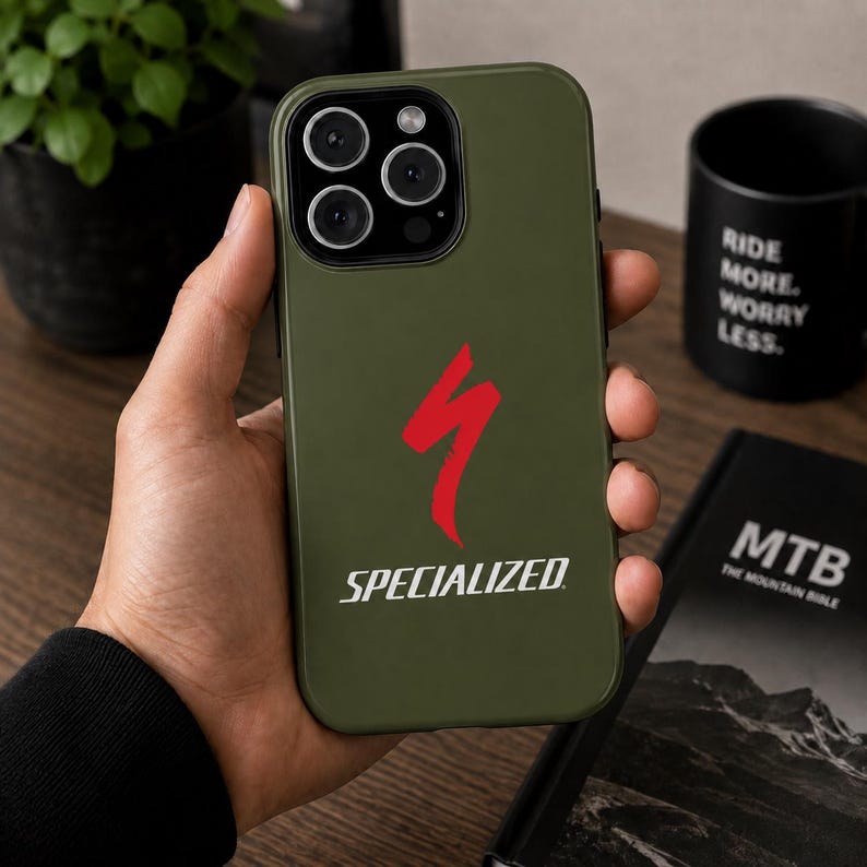 May include: An olive green phone case featuring the Specialized logo in red and the word "SPECIALIZED" in white. The case is held in a hand, with a black mug and a book titled "MTB The Mountain Bible" in the background.