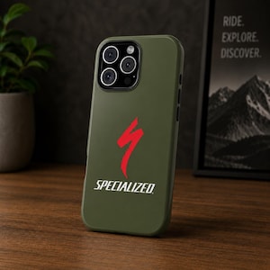 May include: An olive green phone case with the Specialized logo in red and white. The case is on a wooden surface with a plant and a framed print in the background. The text on the print reads "RIDE. EXPLORE. DISCOVER."