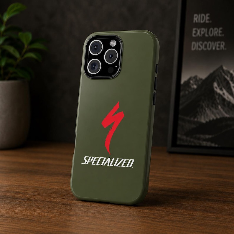May include: An olive green phone case with the Specialized logo in red and white. The case is on a wooden surface with a plant and a framed print in the background. The text on the print reads "RIDE. EXPLORE. DISCOVER."