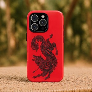 May include: A red phone case with a black illustration of a wolf and a mythical creature. The case has a glossy finish and a camera cutout at the top. The background is blurred, with green and brown tones.