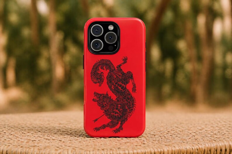May include: A red phone case with a black illustration of a wolf and a mythical creature. The case has a glossy finish and a camera cutout at the top. The background is blurred, with green and brown tones.