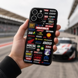 May include: Black phone case featuring numerous racing-related logos, including Mobil 1, HKS, and Brembo. The case has a glossy finish and is held in a hand, with a race car visible in the background. The logos are in various colors and sizes.