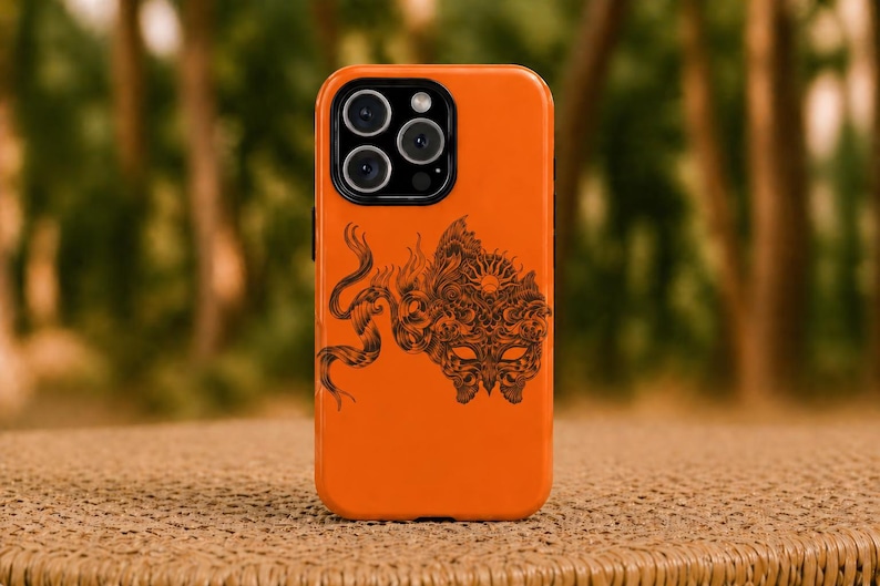 May include: Bright orange phone case with a black graphic design. The design features a detailed illustration of a mask, sun, and a serpent-like creature. The phone case has a glossy finish and a camera bump.