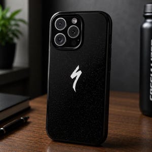 May include: Black phone case featuring a white Specialized logo. The case has a textured finish and a camera module with multiple lenses. The phone case is on a wooden surface.