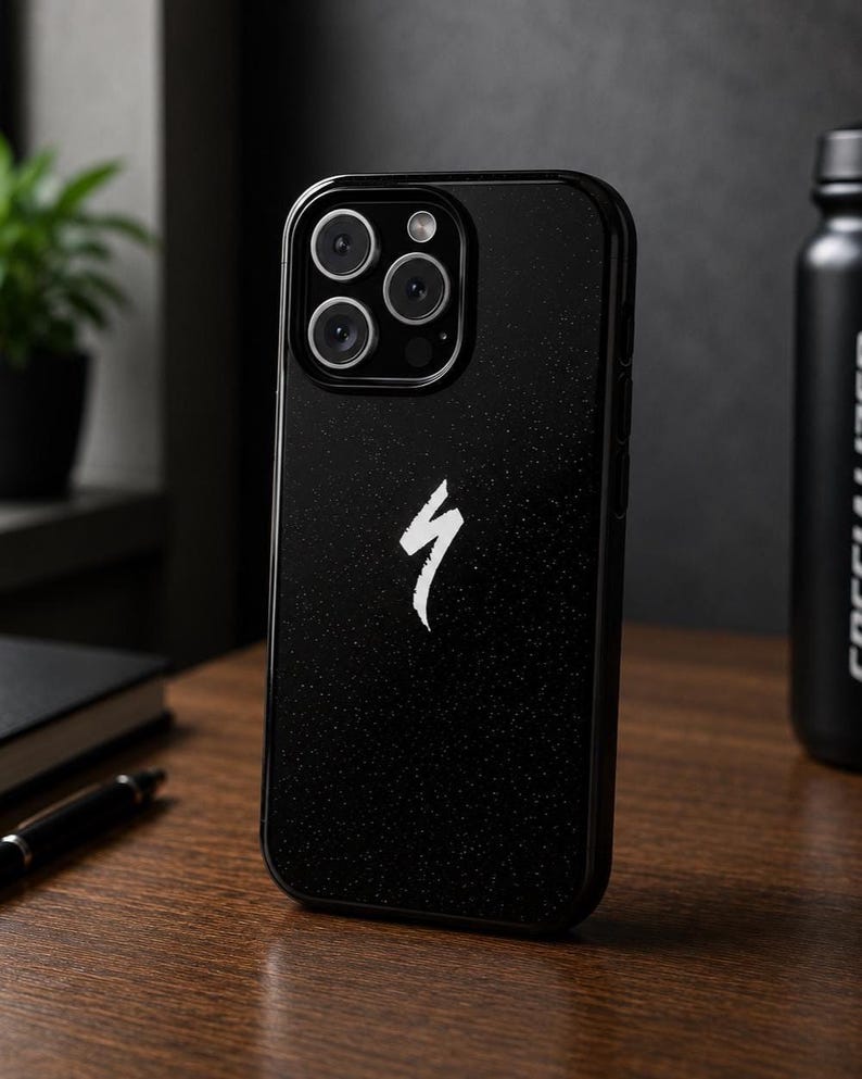 May include: Black phone case featuring a white Specialized logo. The case has a textured finish and a camera module with multiple lenses. The phone case is on a wooden surface.