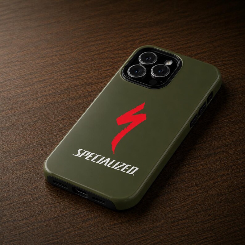 May include: An olive green phone case with a red Specialized logo and the word "SPECIALIZED" in white. The case is on a dark brown wooden surface. The phone case has a camera module with three lenses.