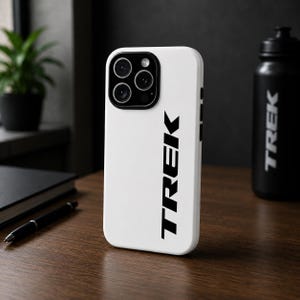 May include: White phone case with the word "TREK" in black, vertical text. The case has a camera module with three lenses. A black water bottle with the word "TREK" is visible in the background.