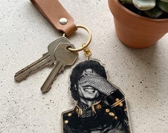 May include: A keychain featuring a black and white image of a smiling person with a hand over their eyes. The keychain has a brown leather strap, gold-colored ring, and two keys.