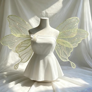 May include: A white strapless top displayed on a mannequin, featuring a small white fabric flower on the left side. Large, translucent, pale green fairy wings with intricate gold glitter veins and scalloped edges are attached to the back.