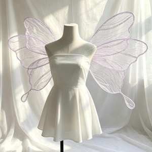May include: A strapless white mini dress displayed on a mannequin, featuring large, translucent, light purple butterfly wings with silver glitter detailing along the edges and veins, extending outwards from the back of the garment.
