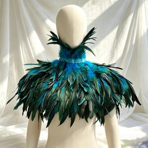 May include: A decorative feather collar in shades of teal and green, presented on a mannequin. The collar has a teal band and a layered design of green and teal feathers. The backdrop is white fabric.
