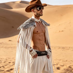 May include: A brown felt cowboy hat, silver-rimmed goggles, and a white cape featuring metallic silver wing-shaped shoulder accents and long white fringe. The outfit includes white fingerless gloves with metal studs and a black studded waist belt.