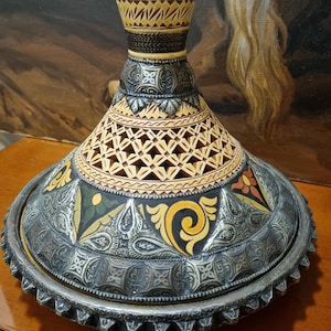 May include: A conical tagine lid featuring a silver-toned metal base with embossed patterns, a central section with intricate geometric cutouts, and a top neck decorated with yellow and black bands, topped with a small silver-toned metal knob.