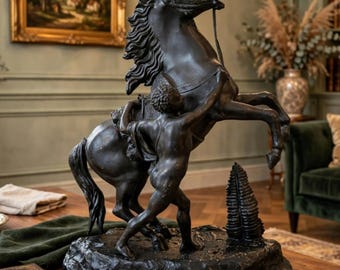May include: A dark bronze-toned sculpture depicting a rearing horse being held by a figure standing beside it. The sculpture is set on a textured, uneven base that includes a small, conical tree-like element. The entire piece is monochromatic.