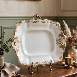 May include: A rectangular white ceramic serving platter with scalloped edges and ornate gold filigree detailing along the border. The platter is displayed upright on a small brass metal stand, featuring a central handle and decorative scrollwork.