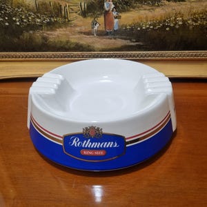 Op de afbeelding: Een ronde witte keramische asbak met een blauwe band rond de basis, voorzien van gouden en rode strepen. De voorzijde toont het Rothmans King Size logo in blauw, goud en rood. De bovenrand bevat vier geïntegreerde rustpunten voor sigaretten.