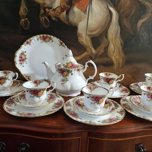 Może przedstawiać: Biały porcelanowy zestaw do herbaty z kwiatowym wzorem czerwonych i żółtych róż, zielonych liści oraz złotymi wykończeniami. Zestaw zawiera dzbanek do herbaty, duży talerz do serwowania oraz cztery filiżanki z pasującymi spodkami i talerzykami.