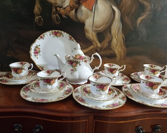 Könnte beinhalten: Ein weißes Porzellan-Teeservice mit einem Blumenmuster aus roten und gelben Rosen, grünen Blättern und Goldrand. Das Set besteht aus einer Teekanne, einem großen Servierteller und vier Teetassen mit passenden Untertassen und kleinen Tellern.