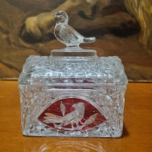 May include: A clear glass trinket box with a bird-shaped lid. The box has a geometric pattern and a red oval design with a bird and leaf motif. The lid features a small, detailed bird figurine.