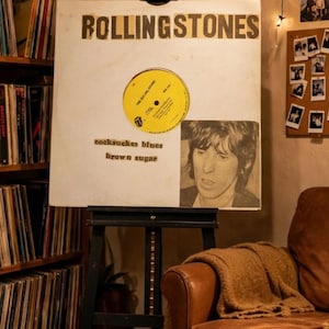 Può includere: Una copertina di disco in vinile bianca con il testo ROLLING STONES in lettere maiuscole nere, un'etichetta circolare gialla e un ritratto in bianco e nero. Il testo cocksucker blues e brown sugar è stampato in nero.