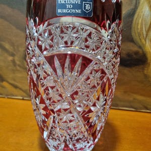 Könnte beinhalten: Eine transparente, geschliffene Glasvase mit tiefrotem Innenraum und Rand. Die Vase weist komplizierte Stern- und geometrische Muster auf. Ein blau-weißer Aufkleber am Rand lautet "EXCLUSIVE TO BURGOYNE".