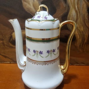 Może przedstawiać: Biały porcelanowy dzbanek do kawy ze złotymi akcentami. Dzbanek ma zakrzywiony uchwyt, długi dzióbek i pokrywkę z małym uchwytem. Ozdobiony złotymi paskami, zielonymi paskami i fioletowymi wzorami kwiatowymi. Stoi na drewnianej powierzchni.