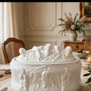 Puede incluir: Una sopera de cerámica blanca con tapa a juego, que presenta intrincados relieves de aves de caza colgadas, tallos de trigo y follaje. El recipiente redondo tiene dos asas laterales y una tapa decorativa con un detalle central.
