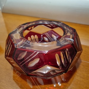 May include: A ruby red, faceted glass bowl with a geometric design. The bowl has a circular opening and is decorated with etched leaf and line patterns. The bowl sits on a wooden surface.