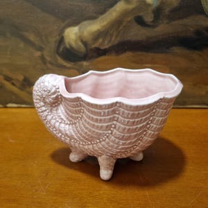 Puede incluir: Una maceta de cerámica rosa claro con forma de concha marina y tres patas pequeñas. La maceta tiene una superficie texturizada con un borde festoneado y probablemente se utiliza para plantas pequeñas o con fines decorativos. El fondo presenta una pintura.