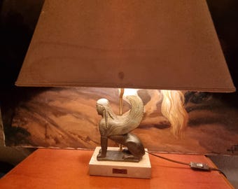 Lampa Le Dauphin Sphinx przedstawia uskrzydloną figurkę stworzenia spoczywającą na jasnej podstawie z trawertynu lub marmuru. Wysokość 50 cm