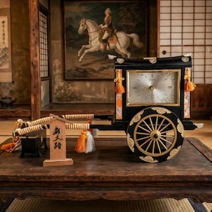 May include: A black and gold decorative clock shaped like a traditional ox-drawn carriage. The clock face is square with silver accents. It features orange and white tassels, gold-toned wheels, and a small wooden sign with black Japanese text.