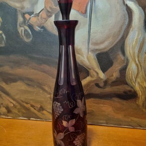 May include: A tall, dark red glass decanter with a matching stopper. The decanter features etched grape and vine designs. The stopper has a rounded top with three holes. The decanter is approximately 10 inches tall.