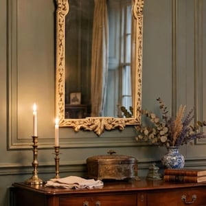 Op de afbeelding: Een boogvormige wandspiegel met een crèmekleurige, rijk bewerkte lijst hangt boven een console-tafel van donker hout. Op de tafel staan twee koperen kandelaars met brandende kaarsen, een metalen doos, een blauw-witte vaas met droogbloemen en boeken.