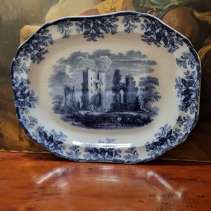 Pode incluir: Uma travessa oval de cerâmica com um padrão de transferência azul e branco. O centro retrata uma paisagem detalhada com ruínas de um castelo de pedra, enquanto a borda recortada é decorada com um motivo repetitivo de folhas de carvalho e bolotas.