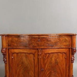 May include: A vintage wooden cabinet with a rich, warm brown finish. The cabinet features two doors and a drawer, ornate carvings, and a curved base. It is mounted on a wooden platform with wheels.