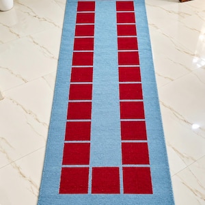 May include: A rectangular runner rug featuring a light blue base color with a repeating pattern of solid red squares arranged in a border around the center. The rug has short, cream-colored fringe tassels along both of the shorter ends.