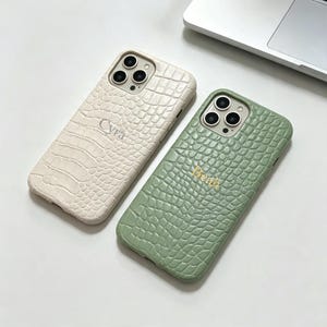 May include: Two smartphone cases with a textured crocodile skin pattern. One case is cream-colored with the name Cyra embossed in the center, and the other is sage green with the name Beah embossed in gold lettering in the center.