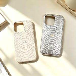 May include: Two rectangular phone cases with a textured snakeskin pattern. The left case is cream-colored with the embossed letters CT, while the right case is metallic silver with the embossed letters MG. Both feature a large camera cutout.