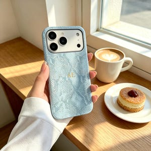 May include: A light blue smartphone case with a textured snakeskin pattern and the gold-coloured initials AE printed in the centre. The case features a rectangular camera cutout with three circular lenses and a flash, covering the back of the device.